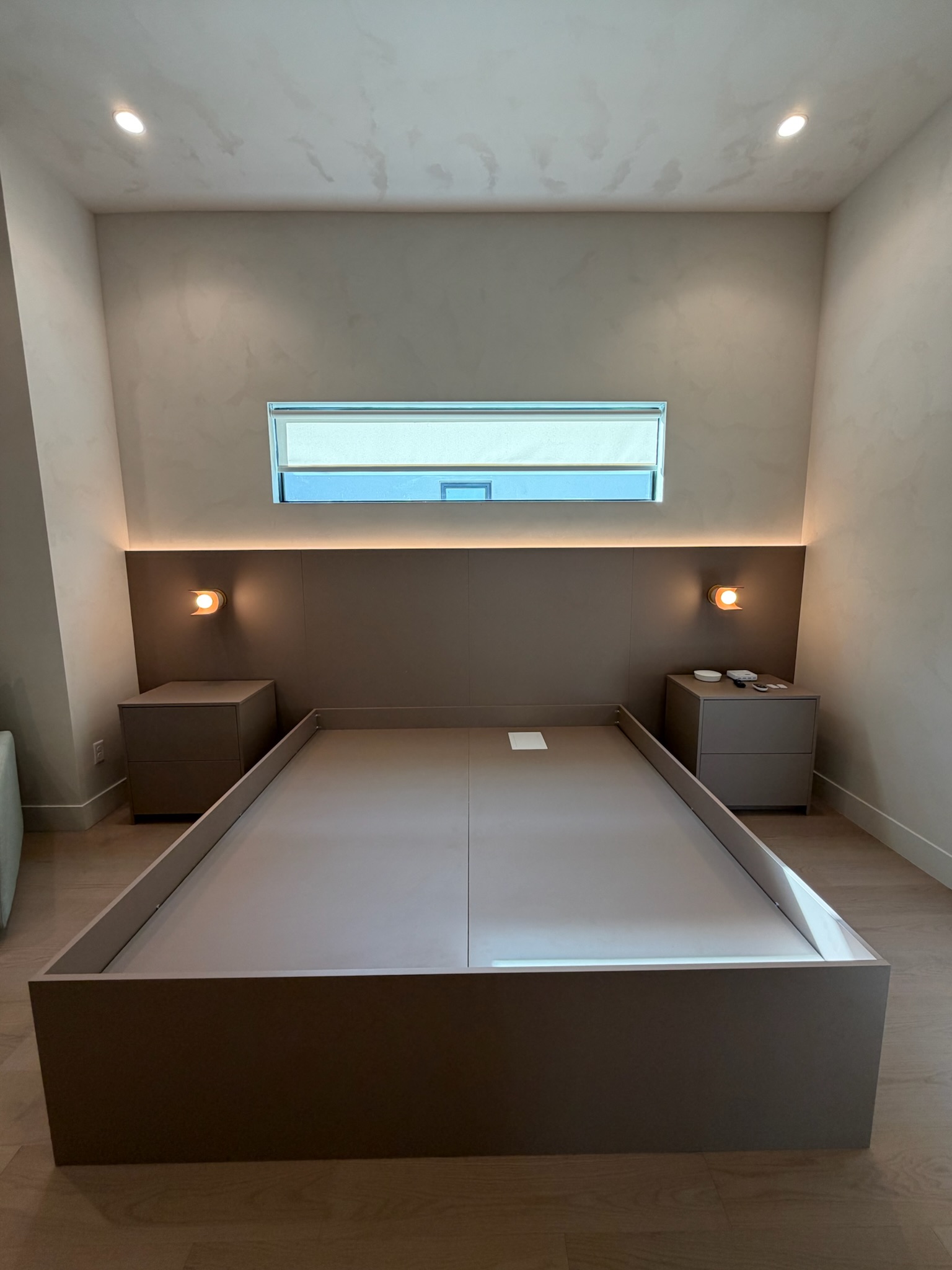 Bedroom with integrated flat-panel platform bed in taupe finish, matching nightstands, and wall-mounted headboard with cove lighting. Minimalist design with concealed storage.