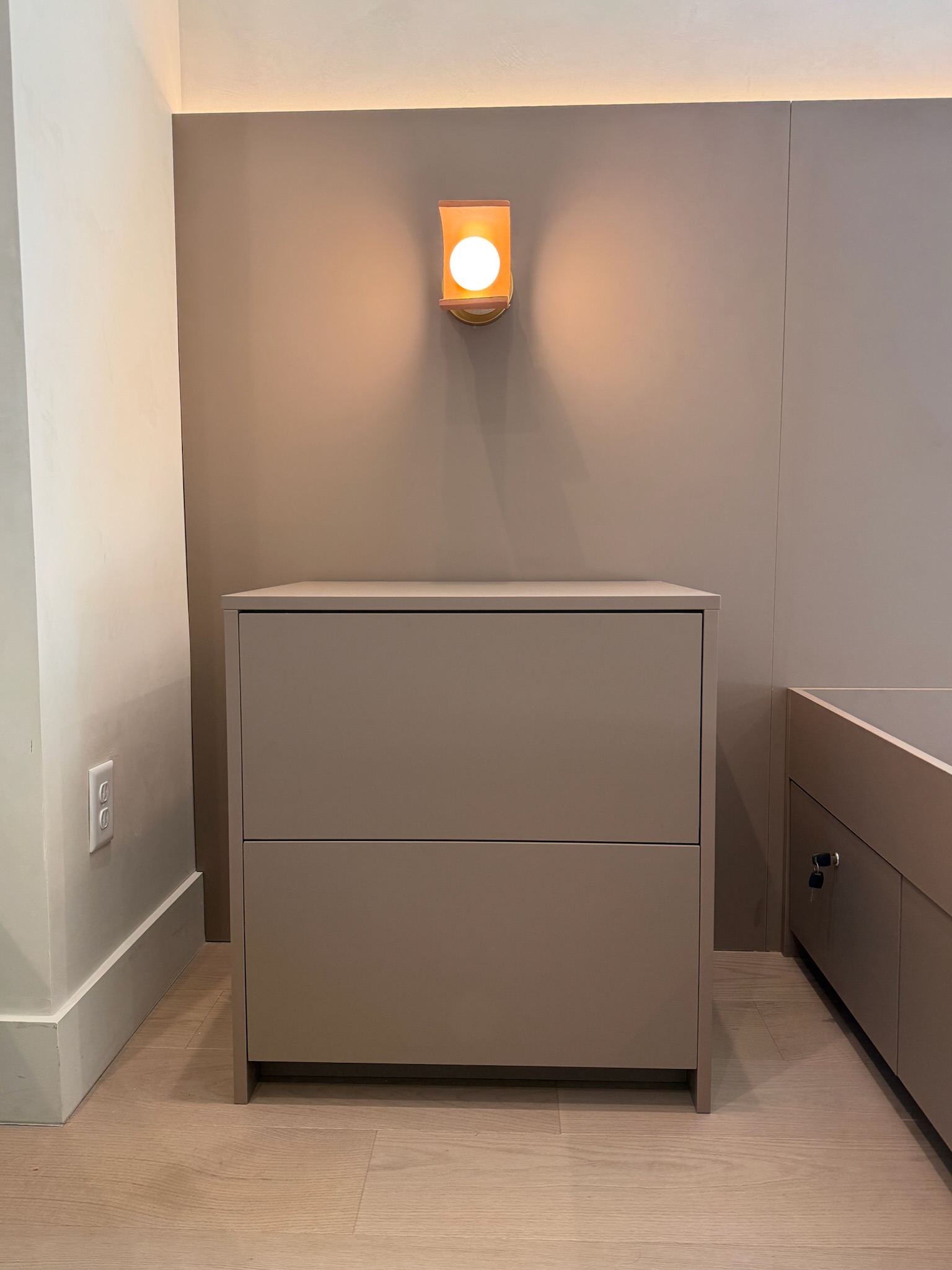 Taupe custom cabinetry with two drawers and integrated toe kick in a bedroom. Recessed wall paneling and wall sconce with orange glass.