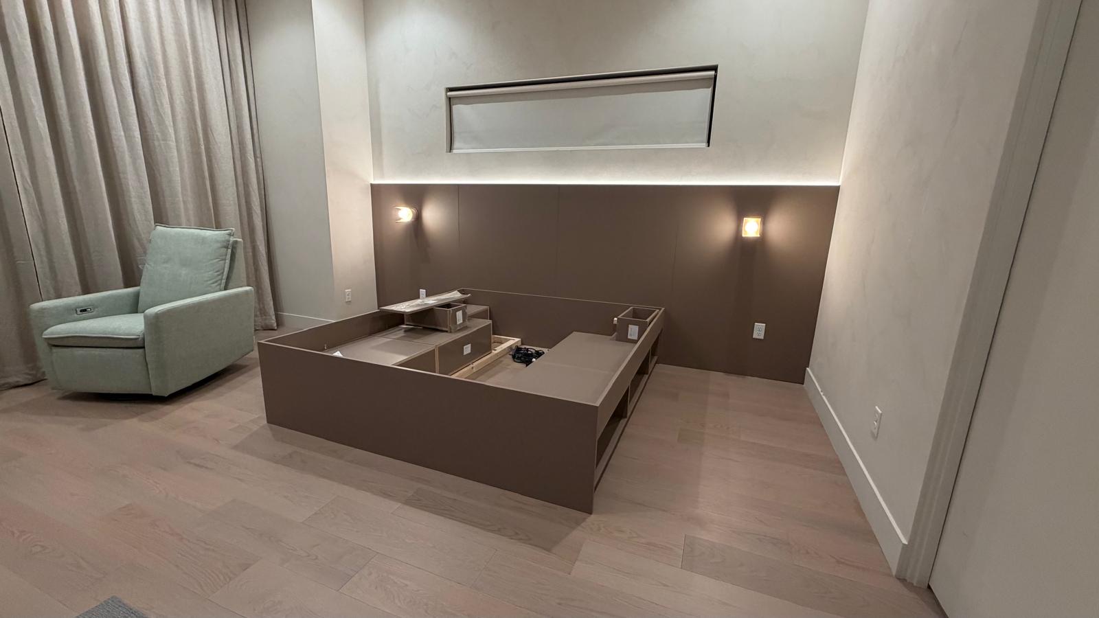 Bedroom with unfinished built-in bed frame and integrated headboard lighting. Light wood flooring, grey green armchair.