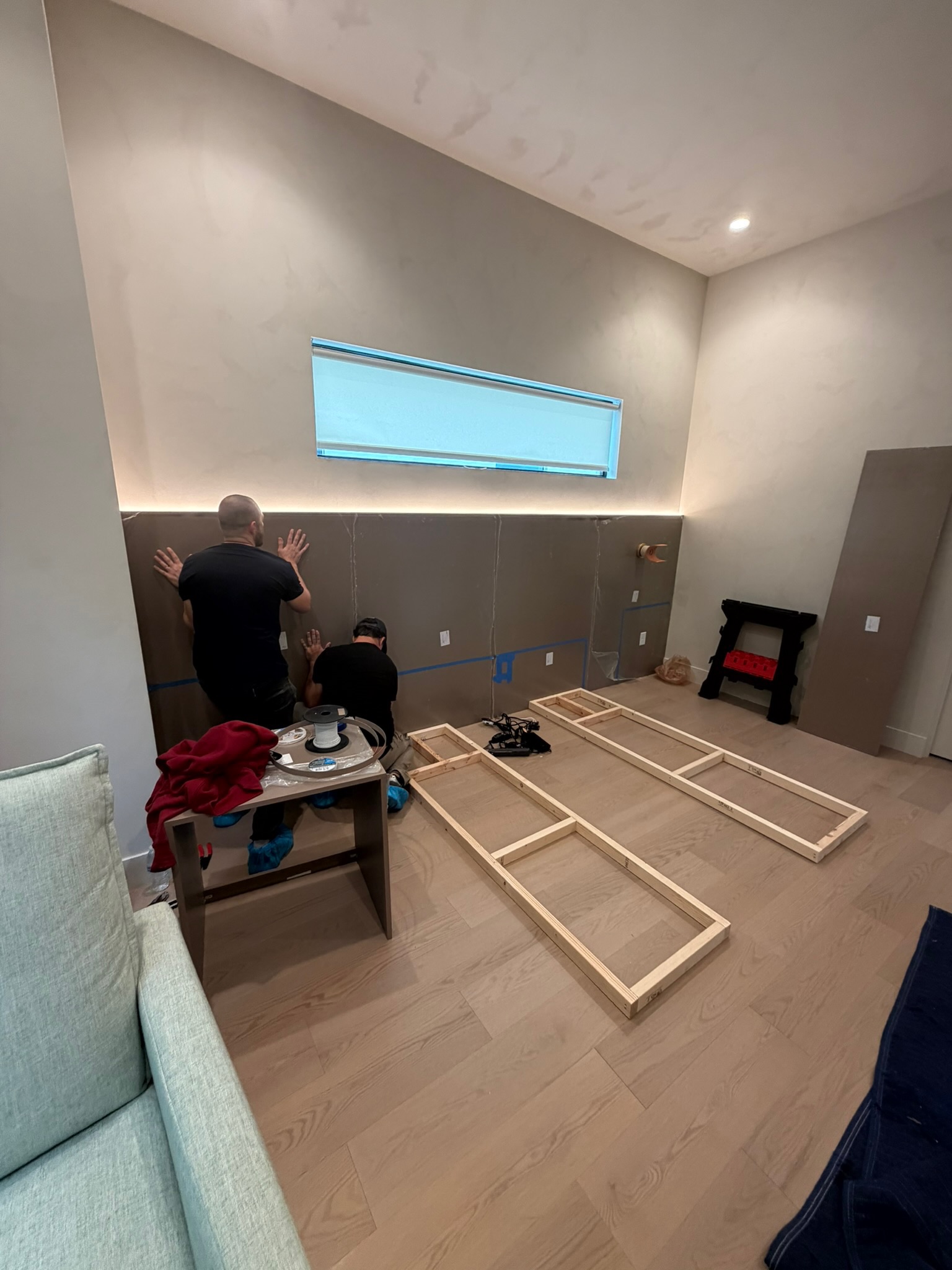 Two workers installing dark wood veneer panels with integrated LED lighting and a long horizontal window in a bedroom.