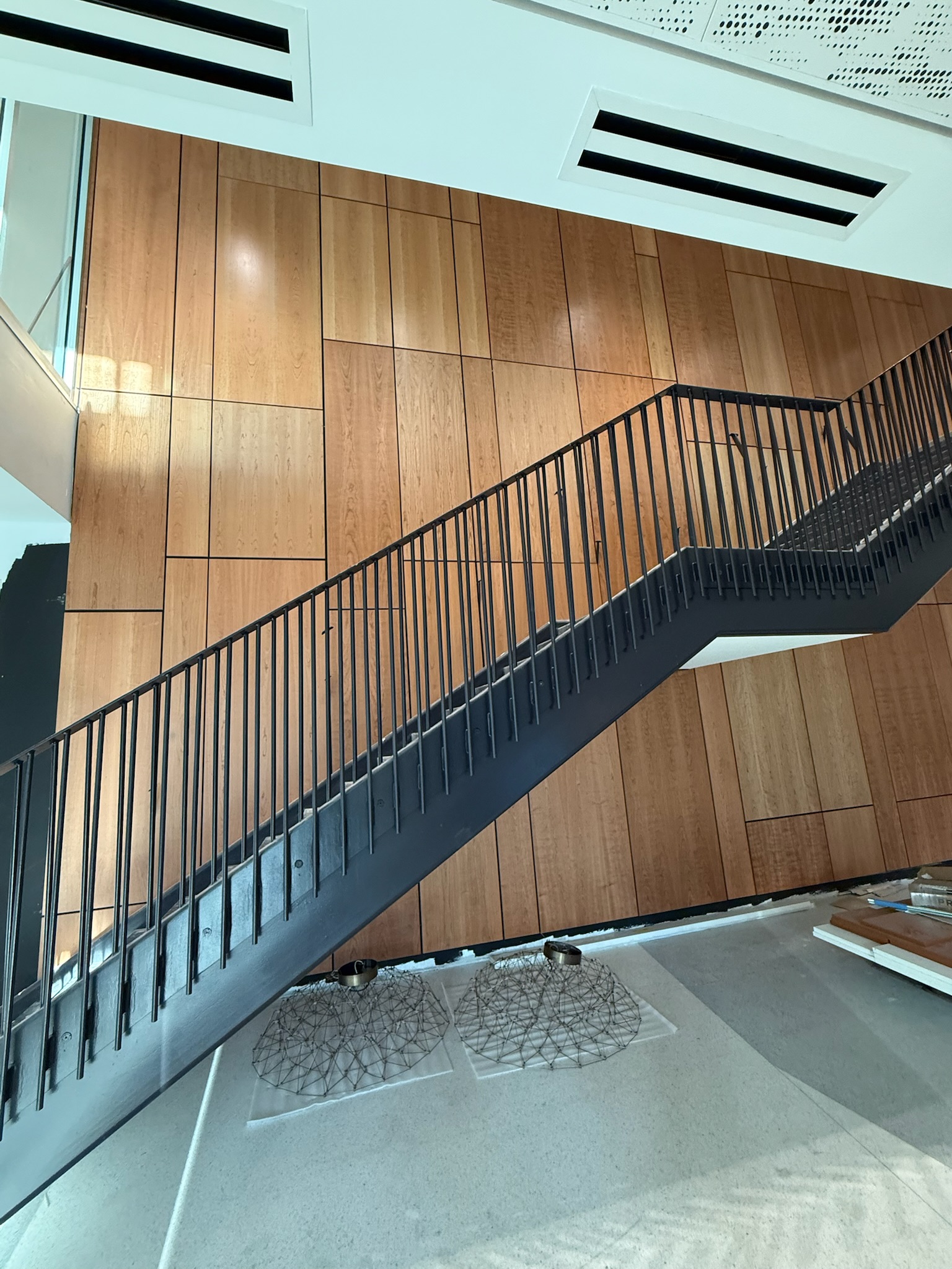 Staircase with black metal railing against a large accent wall of wooden panels in various rectangular sizes, light wood tone, matte finish, commercial building.