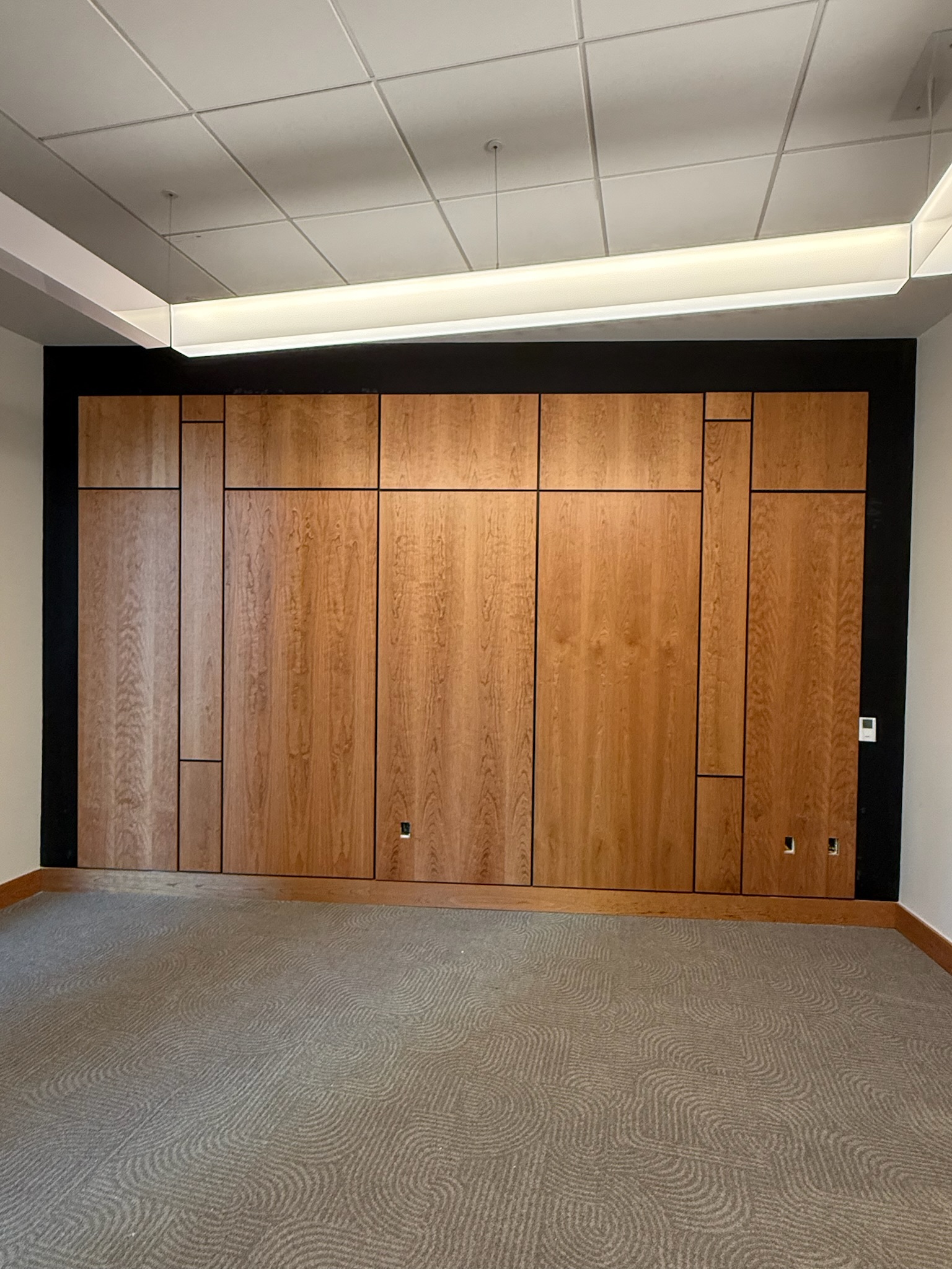 Conference room features wall paneling with varying sized rectangular cherry wood veneer panels, black reveals, and custom base molding, above gray patterned carpet.