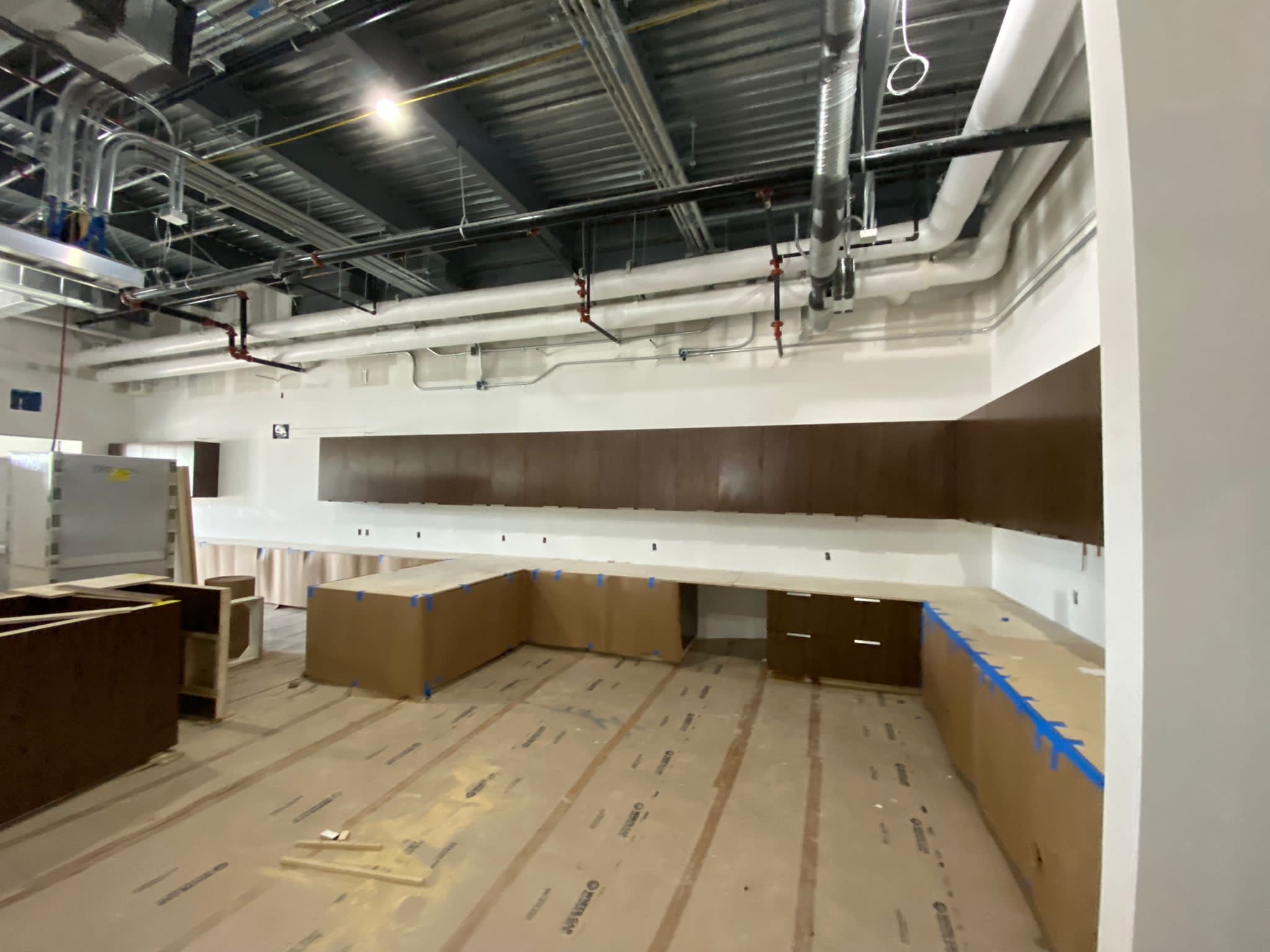 Unfinished food service area with exposed ductwork, dark wood cabinetry, and protective coverings on counters and floors.