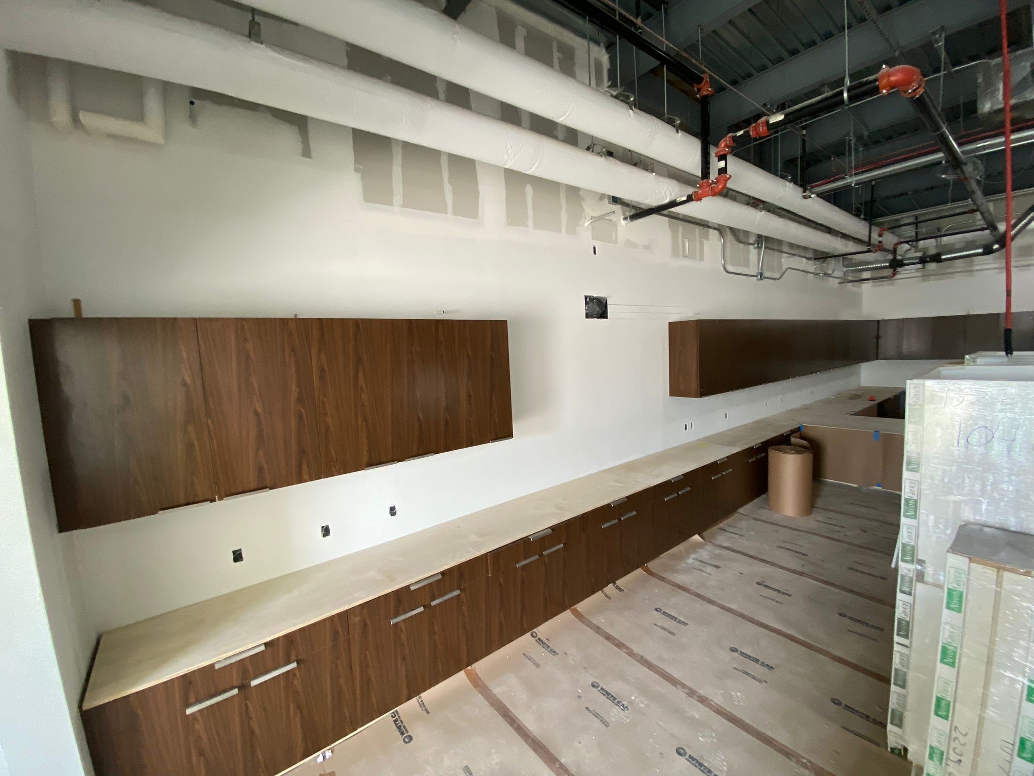 Unfinished luxury stadium lounge with dark wood cabinetry, light countertops, and exposed white and red piping on ceiling.