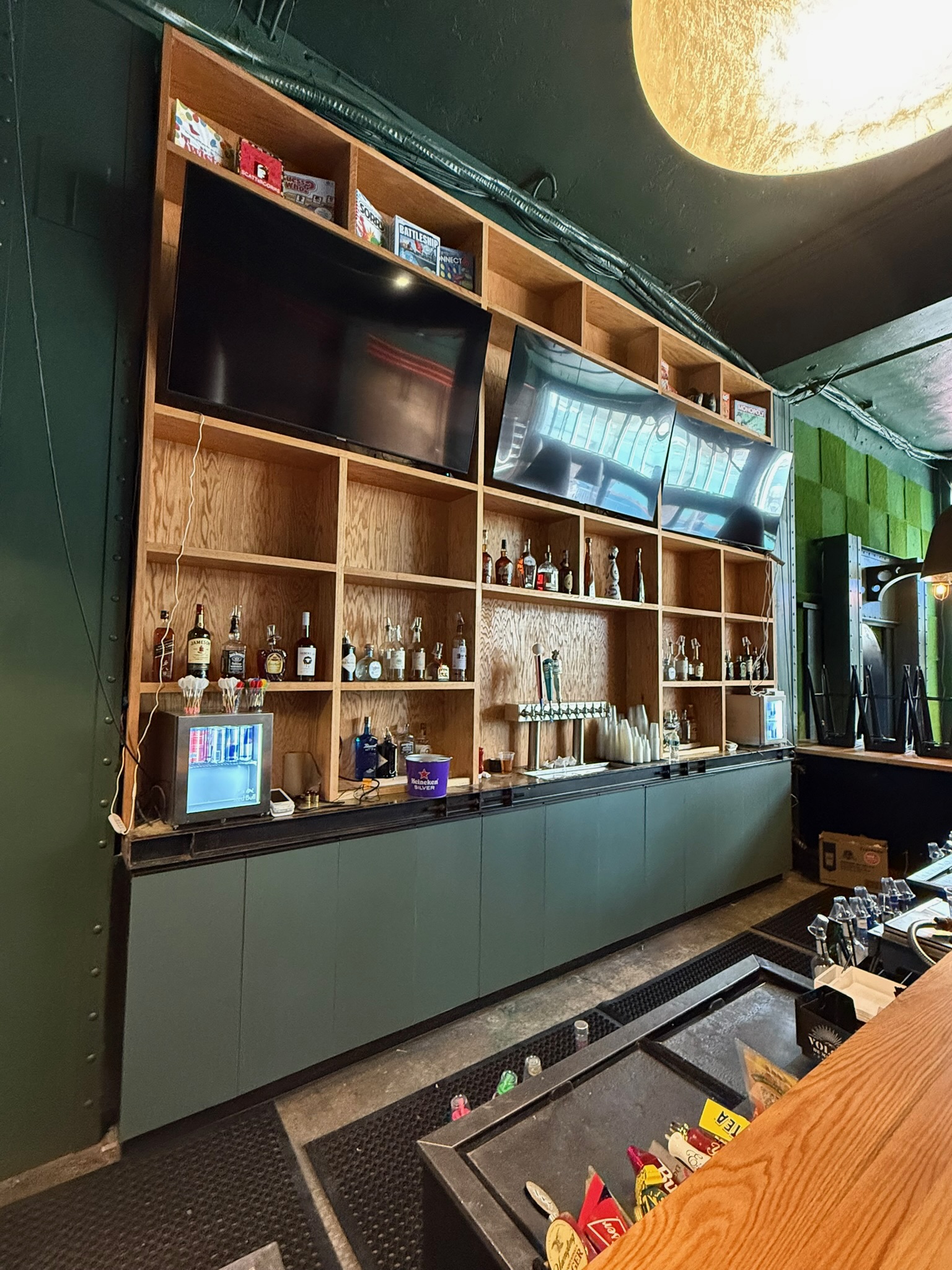 Bar features a custom oak shelving unit with integrated TVs and liquor storage over painted green base cabinets.