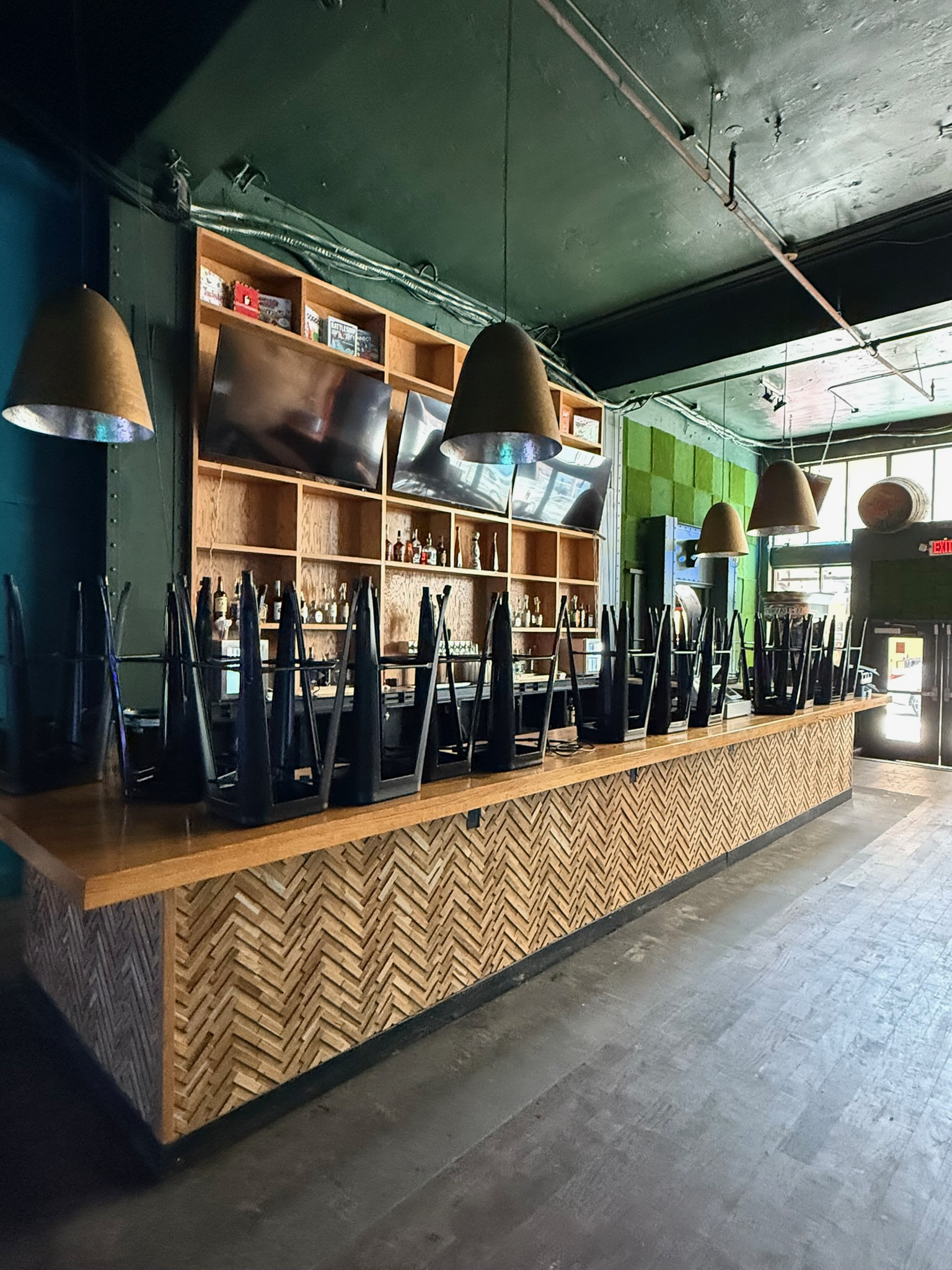 Bar with herringbone wood paneling, wood countertop, open shelving, and black bar stools in a commercial space.