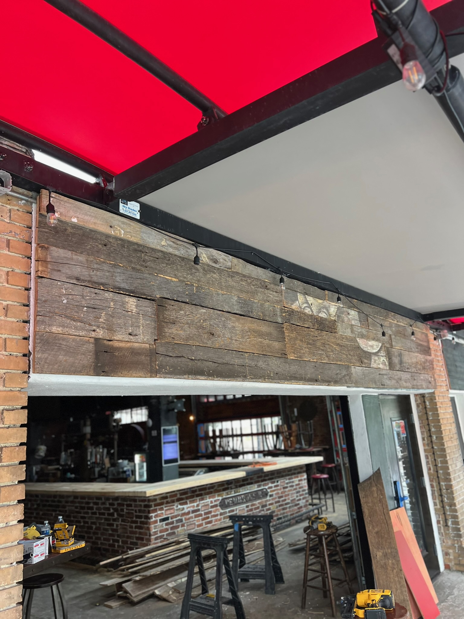 Exterior view of a bar with a red awning. Below the awning is a facade of reclaimed wood planks. The interior shows a brick bar.
