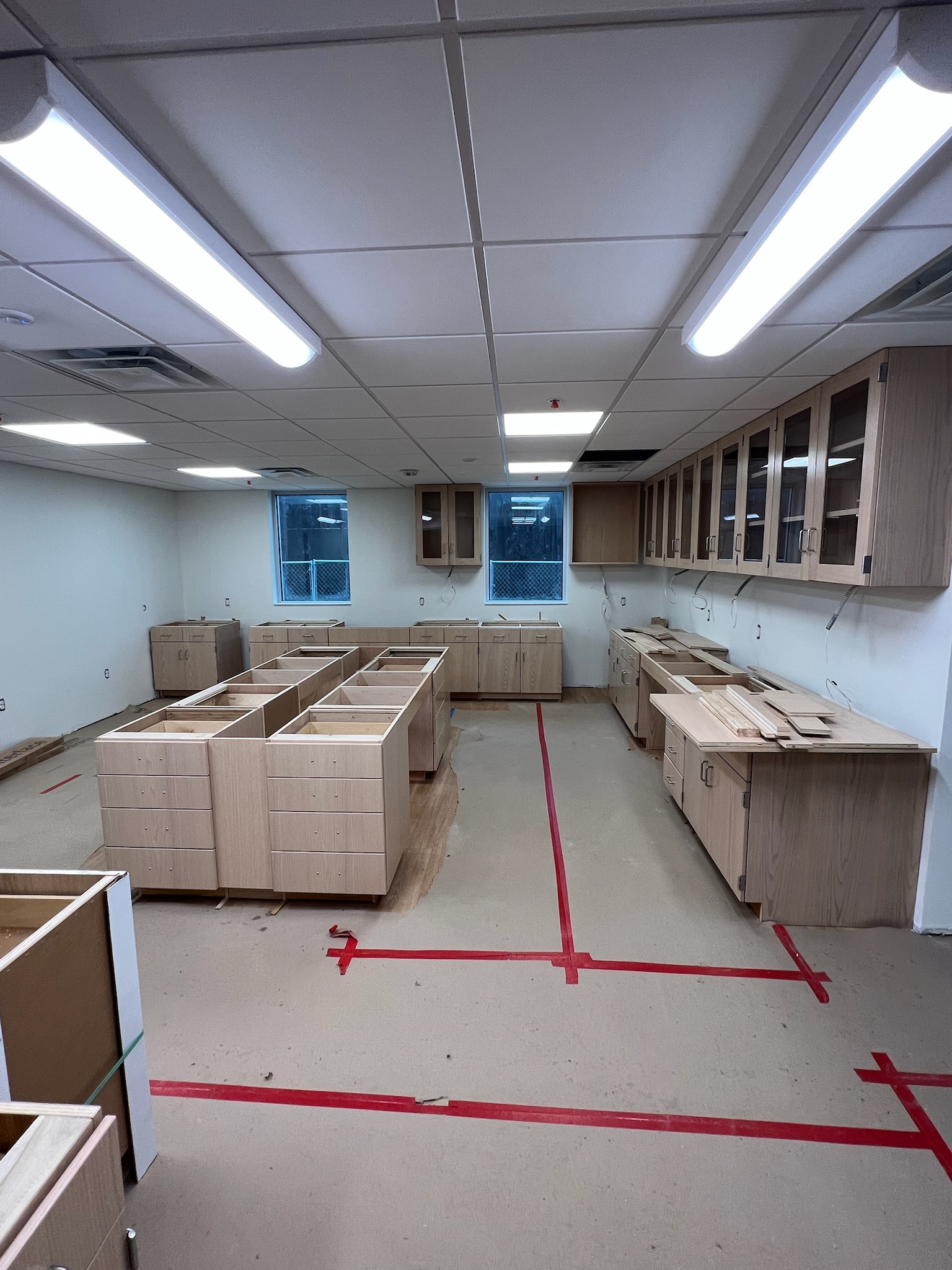 Unfinished birch plywood cabinetry and shelving with glass fronts in a university laboratory. Red tape on floor marks out island and equipment placement.