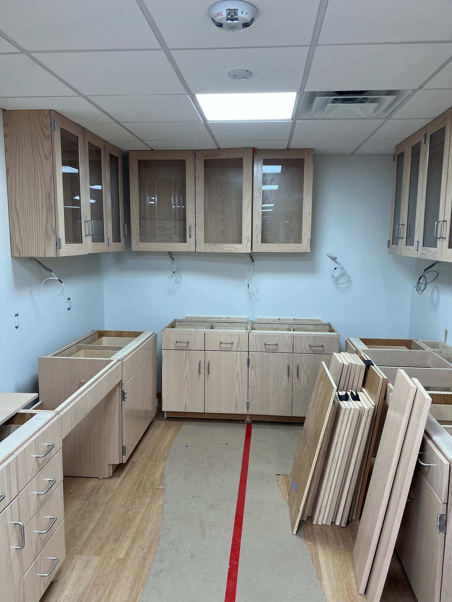 Unfinished laboratory with light wood veneer cabinetry, frosted glass uppers, and stainless steel hardware against white walls with exposed wiring. The dropped ceiling features recessed lighting and a smoke detector.
