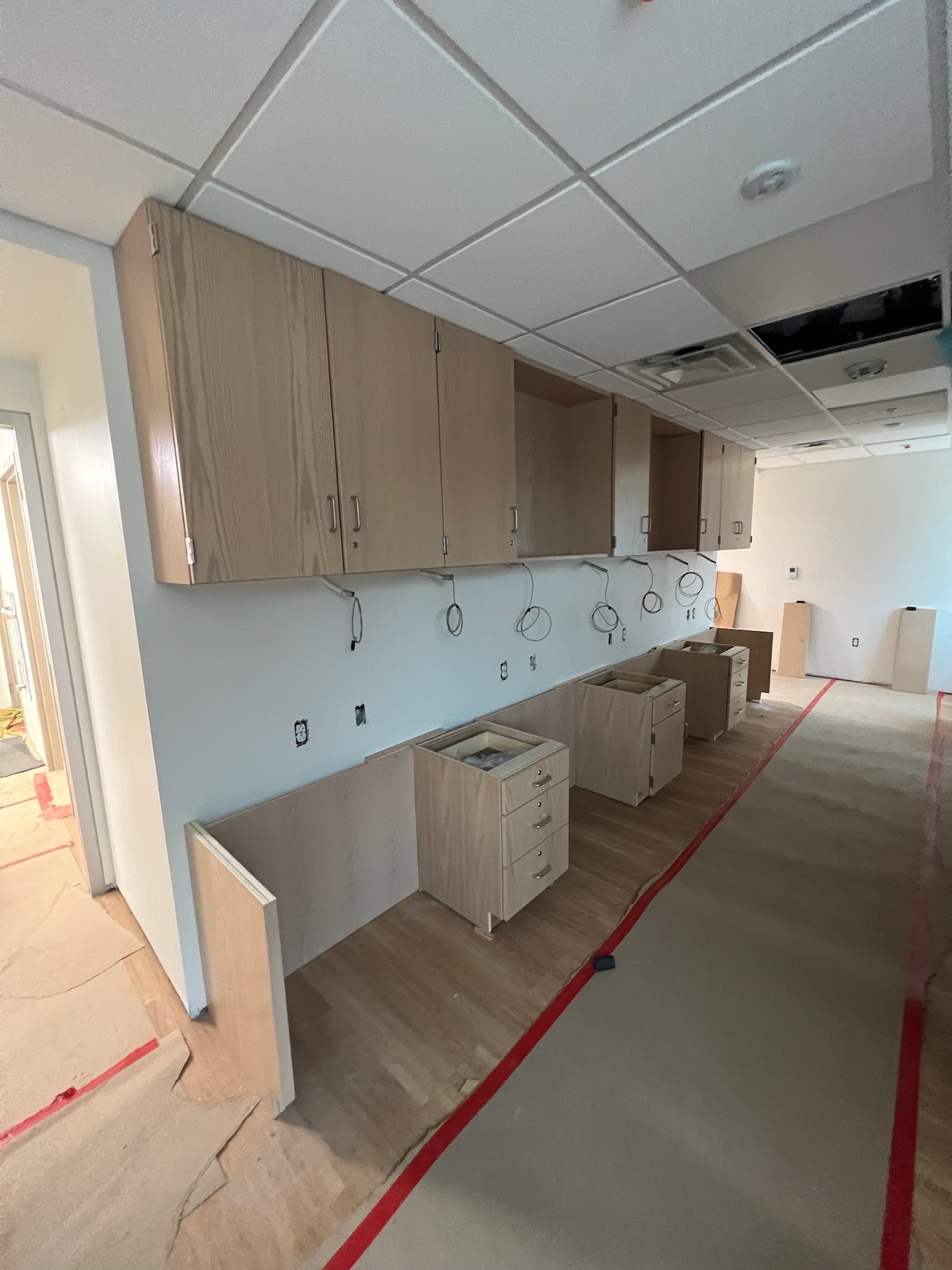Unfinished birch plywood laboratory casework includes upper cabinets and lower drawer units bolted to stud walls and a natural wood floor. Electrical outlets and network cables are visible, awaiting counter installation in a university laboratory.