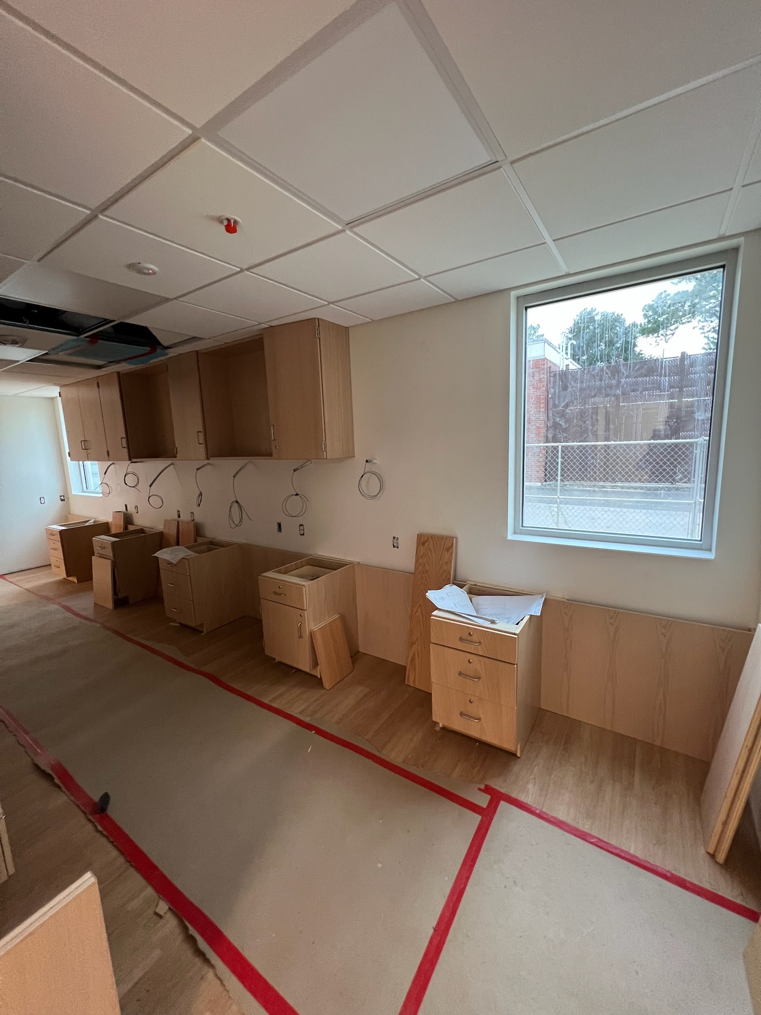 Unfinished birch plywood laboratory cabinets with wall paneling and a drop ceiling. Raw wiring hangs above base cabinets, awaiting countertop installation. A window shows an exterior brick wall and fence.